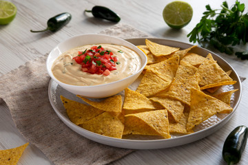 Homemade cheesy dip in a bowl, yellow tortilla chips over white wooden surface, side view. Close-up.