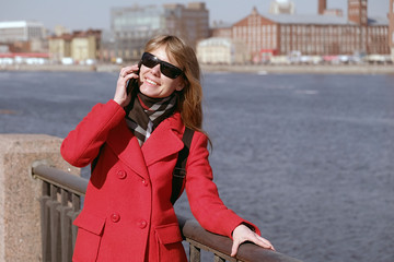 A young girl stands on the waterfront and talking on a mobile phone. Woman happily talking to her friends on the phone and smiling