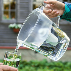 Girl's hand pours clear filtered water from a water filtration jug into a glass which female hand...