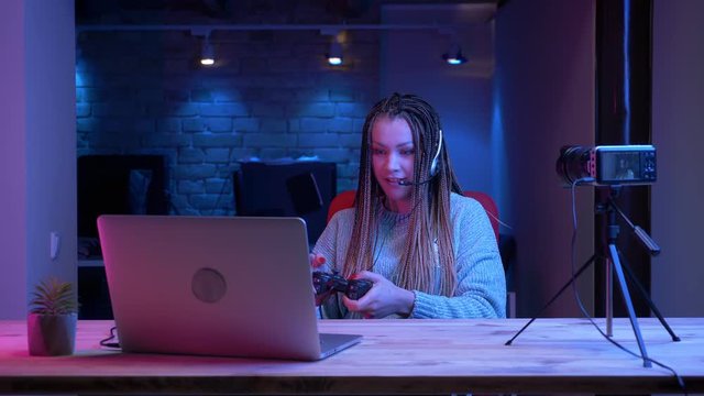 Closeup shoot of young attractive female blogger with dreadlocks in headphones playing video games on the laptop with the neon background indoors