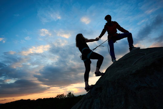 Silhouettes of couple man and woman helping each other climbing up a mountain at sunrise background. Friend helping her. Business, teamwork, success, goal, education, couple, love and help concept.