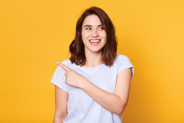 Fototapeta premium Smiling beautiful young woman wears white casual t shiort, points with index finger aside, presenting something and looking away, model poses isolated on yellow background. Copy space for advertisment