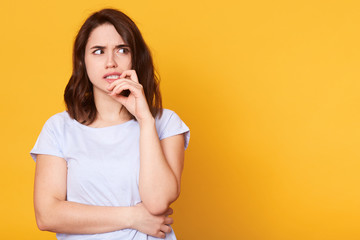 Horizontal portrait of thoughtful female looks with pensive dreamy expression, looking aside, keeps hand near face, thinks what decision to make, stands against yellow background