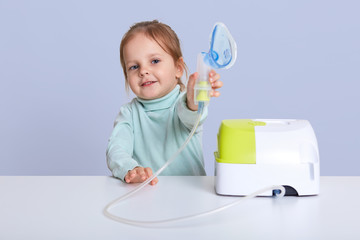 Small attractive girl shows inhaler, makes inhalation with nebulize, sits at white desk, treats...