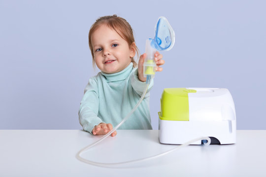 Small attractive girl shows inhaler, makes inhalation with nebulize, sits at white desk, treats illness, dressed casual sweater, posing against blue studio background. Children and health concept.
