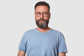 Close up portrait of dark haired serious young male with beard, man looks confidently at camera, dressed casual gray t shirt and round glasses, isolated over white background. People concept.
