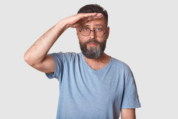 Half length studio shot of handsome male posing isolated over white studio background, peeks out someone, keeps hand near forehead, man dressed casually, has round spectacles, with serious expression.