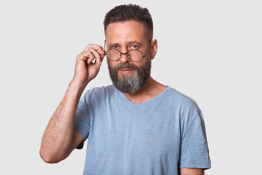 Studio shot of handsome man isolated over white background, looks directly at camera, keeps hand on his spectacles, has stylish hairdo, dressed casual gray t shirt, has serious facial expression.
