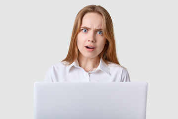 Attractive girl looks at camera with opened mouth, wide opened blue eyes, has straight broun hair, dressed white shirt, working online, shocked with new task, looks astonished, isolated on studio wall