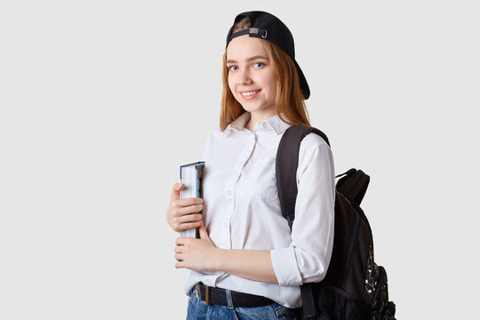 Image of happy Caucasian student with rucksack and big book, dressed shirt, jeans visor cap back, stands smiling isolated over white background, being in good mood, has pleasant facial expression.