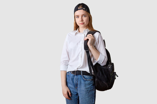 Image of beautiful student girl wearing fashionable jeans nad white shirt, walking to college from home, carrying backpack, looking directly at camera, posing against studio wall with copy space.
