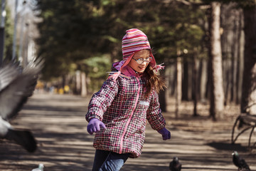 Girl in a red jacket, hat and glasses chasing pigeons in the park. A girl walks along a park alley on a sunny spring day.