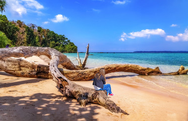Fototapeta na wymiar Young girl tourist sitting on a fallen tree trunk at the scenic sea beach at the Jolly Buoy island at Andaman, India