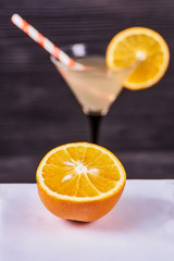Sliced orange next to martini goblet with fresh orange juice and orange slice, cocktail tube, on a dark wooden background. Focus on orange.