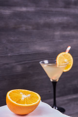 Sliced orange next to martini goblet with fresh orange juice and orange slice, cocktail tube, on a dark wooden background. Focus on orange.