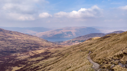 An aerial view of a valley in the Scottish mountain with a lake in the background under a majestic blue sky and white clouds