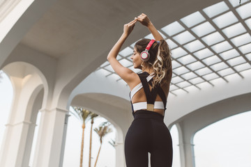 Portrait from back attractive sportswoman with long curly hair in headphones stretching hands above...