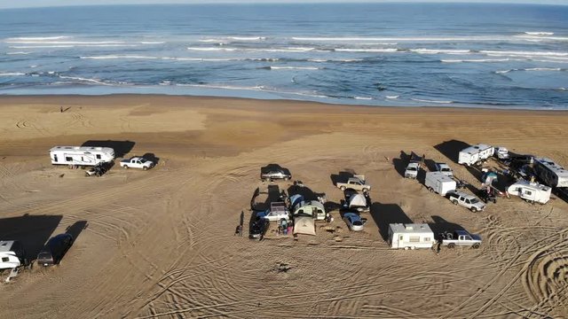 Drone pulls away on the Pismo Dunes with the Ocean in the background while showing many tents, RVs, trucks and campsites on the beach as it flies higher on a bright morning