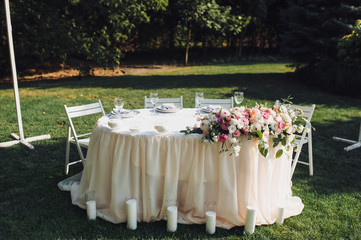 A large white round table, decorated with fresh flowers, candles stands on the nature, in a park with green grass. Wedding decorations and details. Preparing for a wedding party.