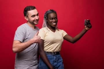 Young mixed race couple, caucasian man and african woman taking selfie from phone while standing on red background