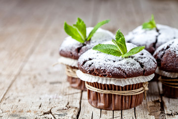 Fresh chocolate dark muffins with sugar powder and mint leaf on rustic wooden table background.