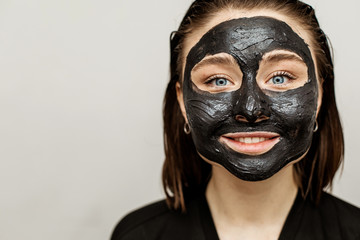 Cheerful nice young woman has black facial mask on face. Smiling and looking straight. Brunette in black bathrobe. Isolated on grey background.