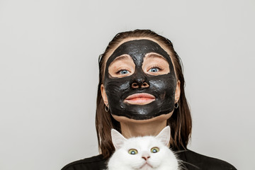 Young woman with black facial mask on skin looking straight. She hold white cat in hands. Wearing black bathrobe. Isolated on grey background.