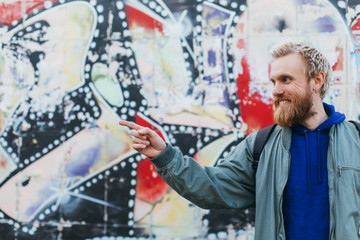 Cheerful nice young man point on wall and smile. Bearde guy stand outisde on street. Urban view.
