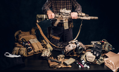Fototapeta na wymiar Man in a checkered shirt holding a assault rifle and showing his military uniform and equipment. Modern special forces equipment. Studio photo against a dark textured wall