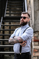 Brutal bearded bully is standing near stairs at slam. He is wearing shirt, suspender and...