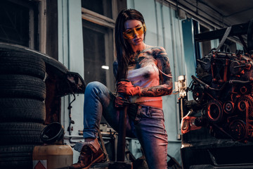 Fototapeta na wymiar Female car mechanic holding a big hammer and posing next to a car engine suspended on a hydraulic hoist in the workshop.