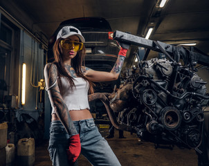 Sexual tattoed girl wearing cap and dirty clothes posing next to a car engine suspended on a hydraulic hoist in the workshop
