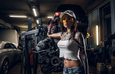 Sexual tattoed girl wearing cap and dirty clothes posing next to a car engine suspended on a hydraulic hoist in the workshop - Powered by Adobe