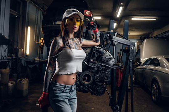 Sexual tattoed girl wearing cap and dirty clothes posing next to a car engine suspended on a hydraulic hoist in the workshop