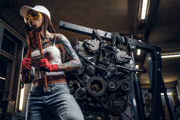 Fototapeta na wymiar Stylish girl wearing cap and goggles posing with a big wrench next to a car engine suspended on a hydraulic hoist in the workshop