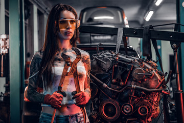 Obraz na płótnie Canvas Female model with tattooed body wearing protective goggles posing with a big wrench next to a car engine suspended on a hydraulic hoist in the workshop. Photo with red light illumination