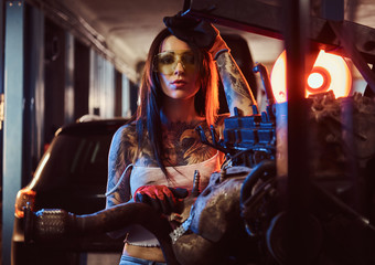Fototapeta na wymiar Beautiful female model with tattoed body repairs a car engine suspended on a hydraulic hoist in the workshop. Photo with red light illumination