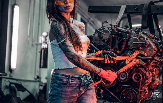 Cropped image. Stylish female model with tattoed body repairs a car engine suspended on a hydraulic hoist in the workshop. Photo with red light illumination 