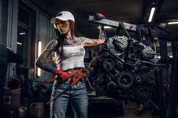 Plakat Female model with sexy tattooed body wearing a cap and protective goggles posing with a big wrench next to a car engine suspended on a hydraulic hoist in the workshop .