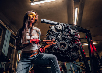 Fototapeta na wymiar Stylish female model with tattoed body repairs a car engine suspended on a hydraulic hoist in the workshop.