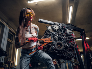 Obraz na płótnie Canvas Stylish female model with tattoed body repairs a car engine suspended on a hydraulic hoist in the workshop.