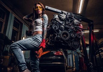 Fototapeta na wymiar Stylish tattooed girl holding a big wrench and posing next to a car engine suspended on a hydraulic hoist in the workshop.