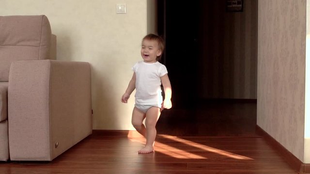 Little cute baby girl makes the first independent steps at home and laughs merrily in slow motion without any help . Kid begins to walk confidently on his feet. One year old girl learning to walk