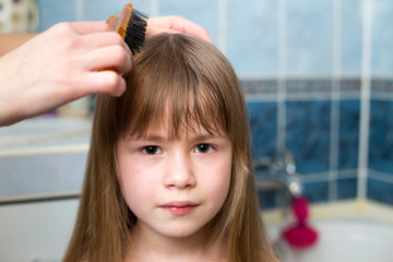Fototapeta premium Mother hand with brush combing long fair hair of cute child girl after bath on blurred interior background.