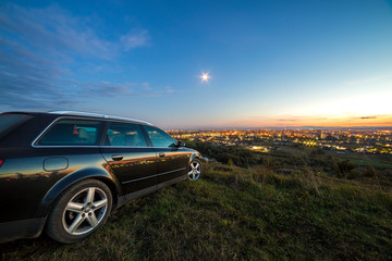 Black car parked at night in green meadows on copy space background of lights of distant city buildings and bright blue sky with first star at sunset.
