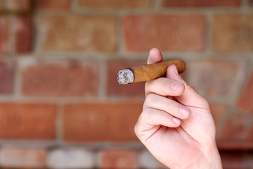 A young man holds a burning and cigar in his hand with smoke. Smoker and smoking with brick wall on...