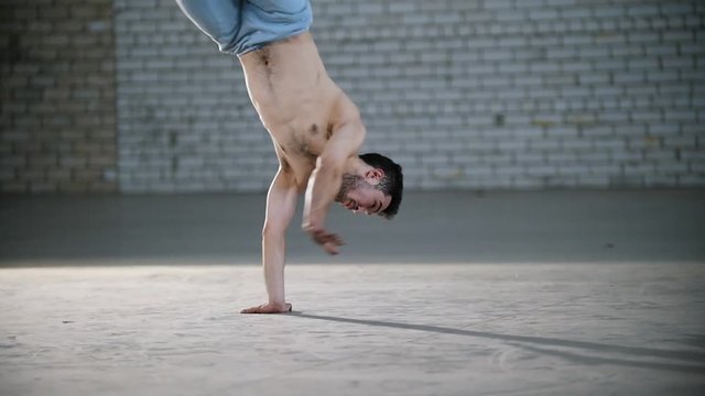 A bright room. An athletic shirtless man training his capoeira skills and standing on his hands