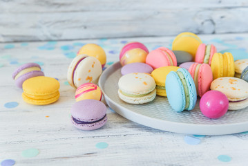 Fototapeta na wymiar Multicolored french macaron cakes on blue plate and white wooden background. White, yellow, pink, purple and grey french macarons with fresh berries.