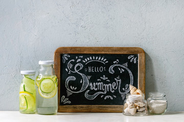 Summer theme. Chalk lettering Hello summer on vintage chalkboard, seashells and sand in glass jars, sunglasses, two glass bottles with sassy water on white marble table, concrete wall at background.
