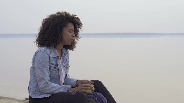 Positive, happy afro american woman outdoors. Sitiing near the lake with cup of coffee (oe tea), singing, enjoying life.